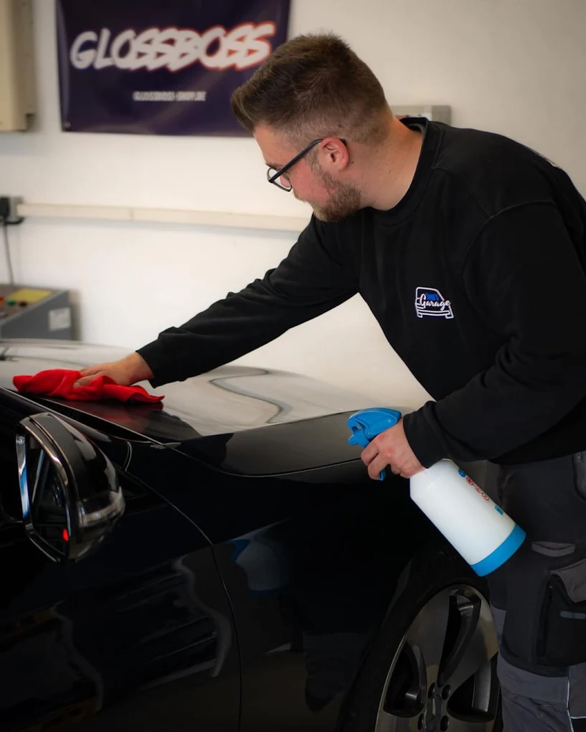 Simons Garage - Fahrzeugaufbereitung - Simon May reinigt ein schwarzes Auto mit einer Sprühflasche und einem roten Tuch.